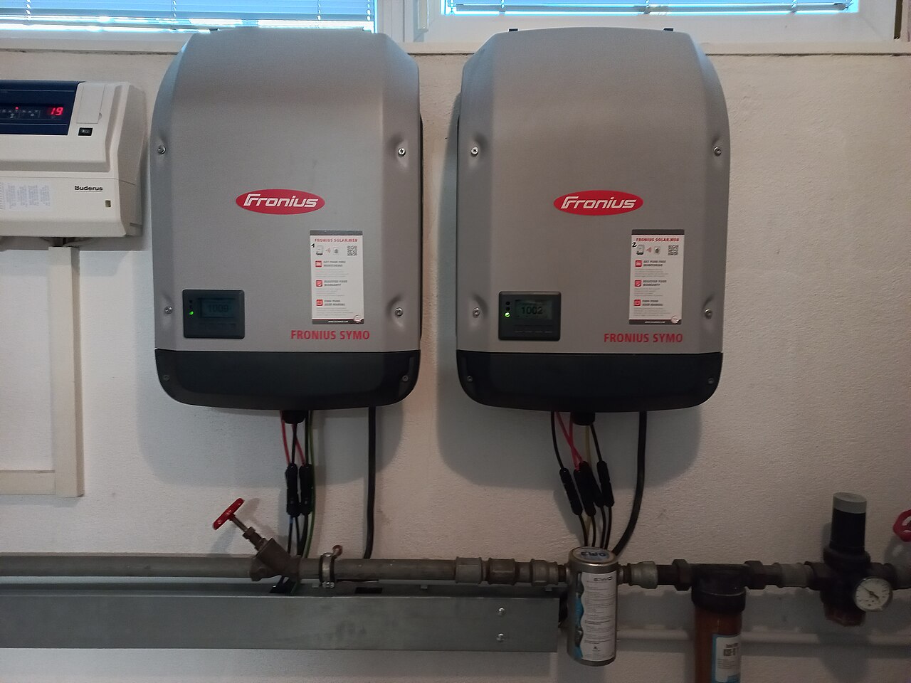 Two grey Fronius Symo inverters, the three-phase sibling of the Primo in the same SnapINverter housing, mounted side by side on a wall above water pipes
