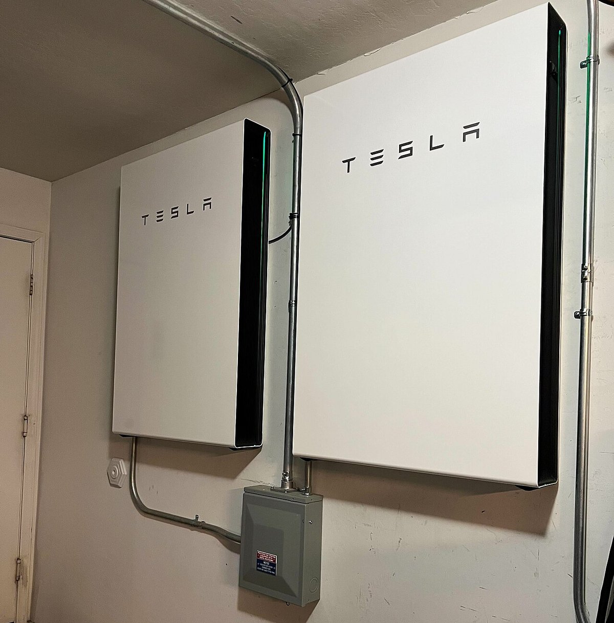 Two white Tesla Powerwall 3 units mounted on an indoor garage wall above a grey disconnect box, photographed in a US home